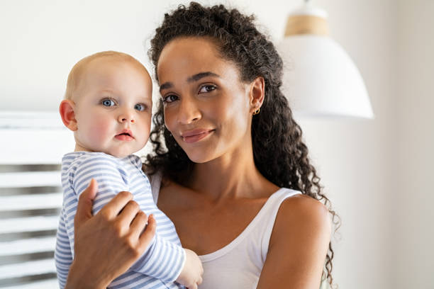 Multiethnic young mother holding cute little baby Smiling african mother holding adopted baby at home. Portrait of proud young woman rocking infant while looking at camera. Beautiful mixed race babysitter carrying adorable little child at home. biracial newborn stock pictures, royalty-free photos & images