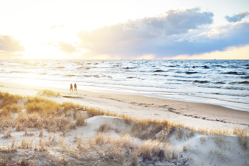 A couple is walking through the Baltic sea shore at sunset. Sand dunes and plants close-up. Colorful evening clouds. Waves and water splashes. Idyllic seascape. Latvia