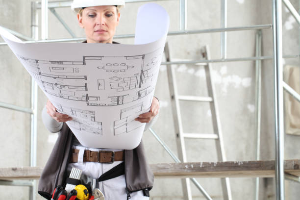 istock woman construction worker builder looking at bluprint, wearing helmet, hearing protection headphones and tools belt bag in building site indoors background 1219127749