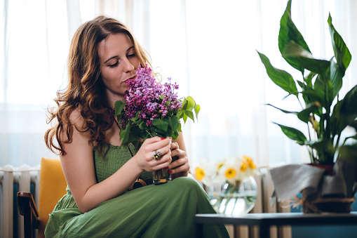 Young beautiful Caucasian woman smelling bouquet of lilac in her living room.