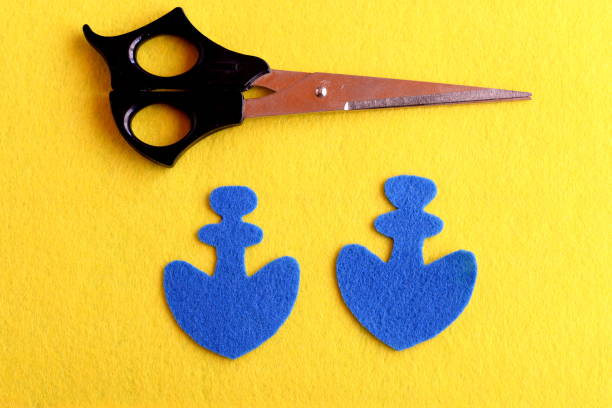 istock Anchor shaped details cut from felt. Two blue felt anchors, scissors isolated on a yellow background. Sewing hobby for children, women, beginners. DIY keychain step by step. Top view 1219135912