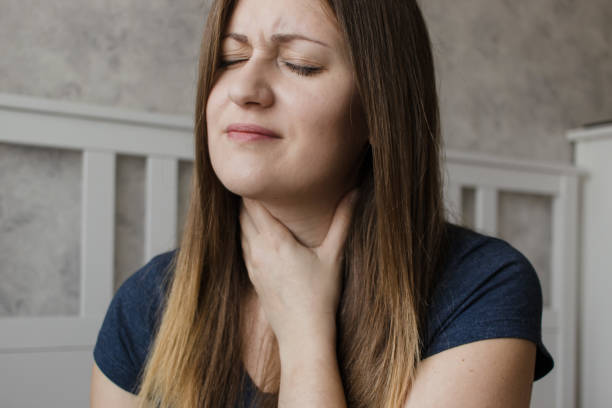 young unhappy woman has sore throat. the girl feels pain, symptoms of the beginning of a cold, flu or virus. tired girl sitting at home having pain in the neck - touching neck imagens e fotografias de stock