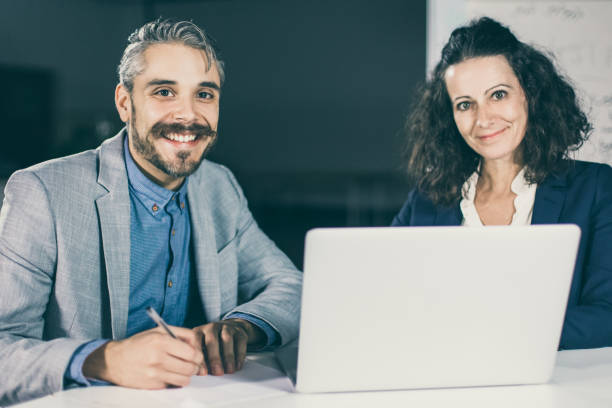 istock Smiling colleagues sitting at table and looking at camera 1219139137