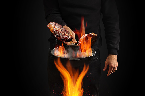 Young male dressed in jumper and jeans. He is holding burning wok pan with smoke above fire and frying two beef steaks, against black studio background. Cooking concept. Close up, copy space