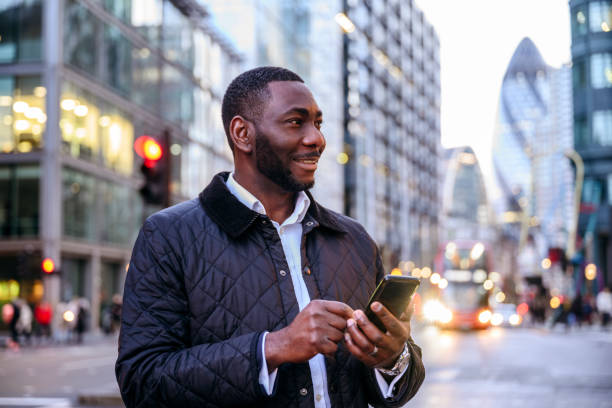 Portrait of African businessman outdoors with smart phone Three-quarter view of smiling London businessman wearing waxed jacket and looking away from camera while checking smart phone outdoors at dusk. businessman african descent on the phone business person stock pictures, royalty-free photos & images