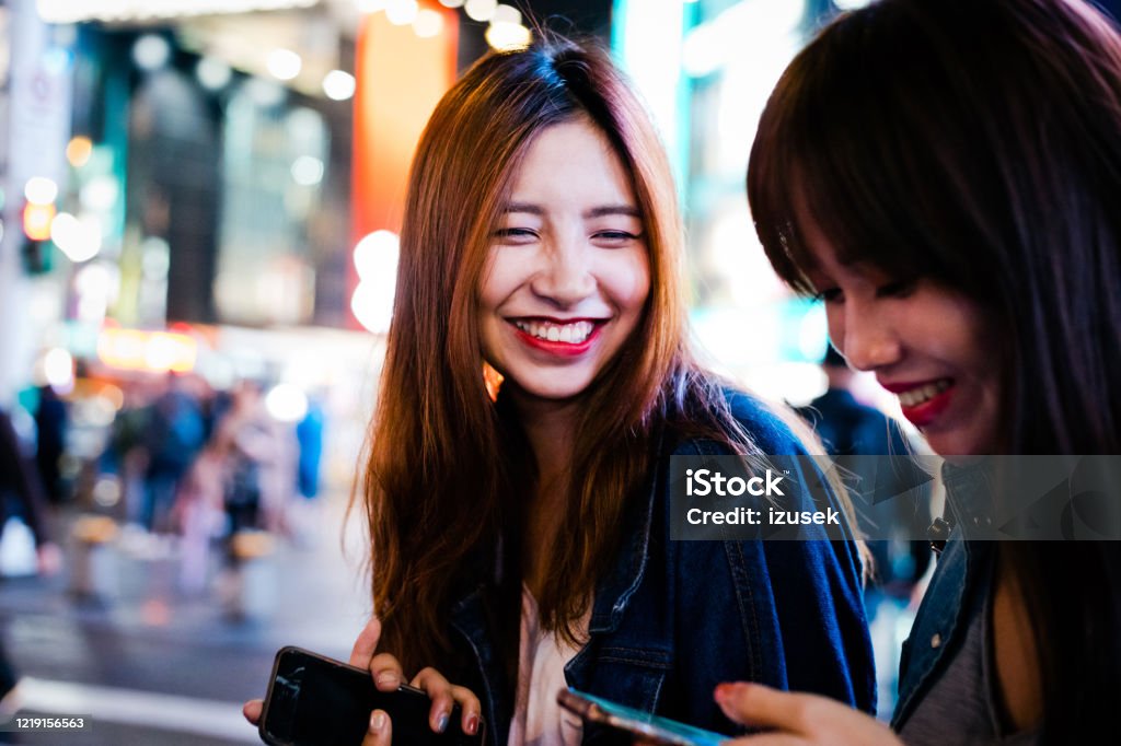 Young women using smart phones on city street at night Female friends using smart phone while standing on street. Beautiful young women enjoying weekend in city at night. They are wearing casuals. 20-24 Years Stock Photo