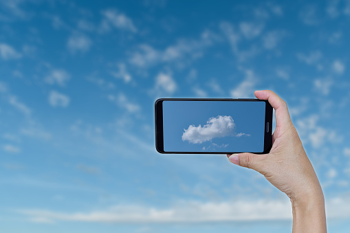 Hand holding smartphone for take photo a single cloud over blue sky background.