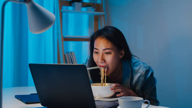 asia freelance smart business donne che mangiano spaghetti istantanei mentre lavorano su laptop in soggiorno a casa di notte. - japanese ethnicity women asian and indian ethnicities female foto e immagini stock