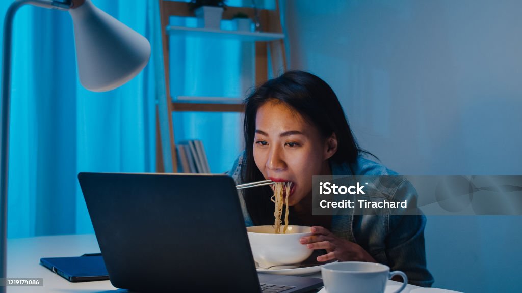 Asia freelance smart business women eating instant noodles while working on laptop in living room at home at night. Asia freelance smart business women eating instant noodles while working on laptop in living room at home at night. Happy young Asian girl sitting on desk work overtime, enjoy relax time. Eating Stock Photo