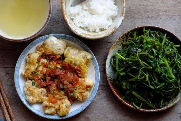 Simple Vietnamese vegan meal for lunch, boiled water spinach, stuffed cabbage with tomato sauce, bowl of rice on wooden background, healthy food that rich fiber