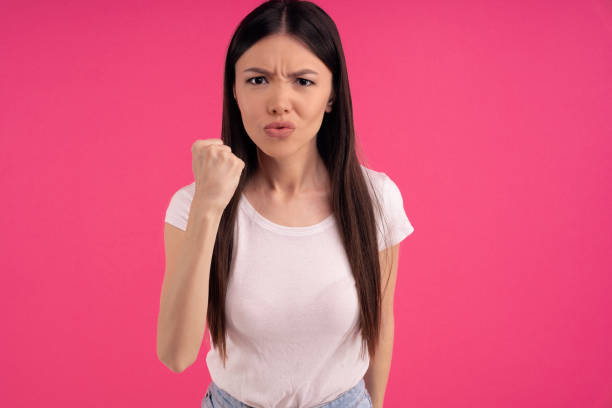 Picture of angry dissatisfied Asian woman clenches fist with displeasure, keeps lips folded, makes outraged face expression, wears white clothing, threatens you, isolated on pink background. Picture of angry dissatisfied Asian woman clenches fist with displeasure, keeps lips folded, makes outraged face expression, wears white clothing, threatens you, isolated on pink background revenge stock pictures, royalty-free photos & images