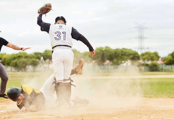 Intense baseball action: A player in a green uniform slides into a base, kicking up dirt, while a catcher in blue and white, captured with a three-quarter length portrait, gear prepares to tag him. The scene captures the dynamic energy of the sport on a sunny field, highlighting athleticism and competitive spirit.