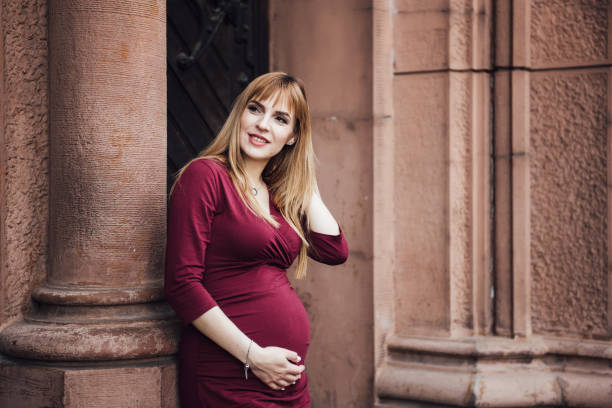 Pregnant girl blonde in burgundy dress. In the big city. Long hair. 9 months waiting. Happiness to be a mother. Against the backdrop of a beautiful old building. Pregnant girl blonde in burgundy dress. In the big city. Long hair. 9 months waiting. Happiness to be a mother. Against the backdrop of a beautiful old building. 8 months pregnant stock pictures, royalty-free photos & images