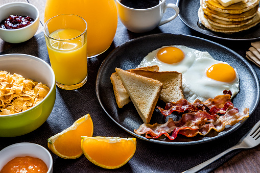 High angle view of an American breakfast made with fried eggs, toasted bread, bacon, pancakes, orange juice, coffee, and oranges.