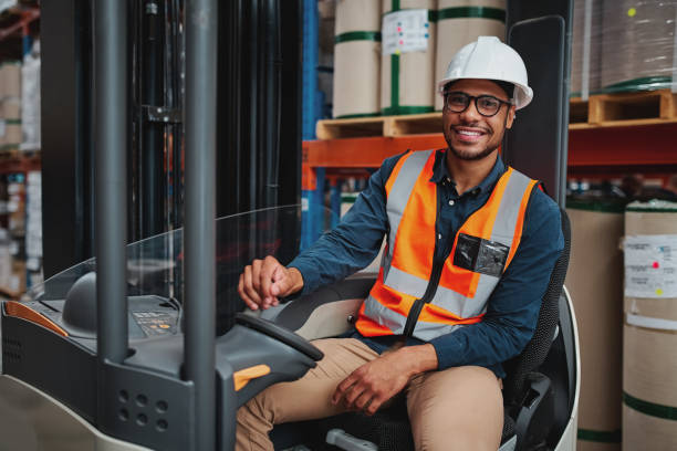 istock Smiling forklift driver with spectacles sitting in vehicle in warehouse looking at camera wearing a white helmet and safety vest 1219190983