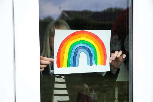 Mujer mayor sosteniendo el dibujo del arco iris en la ventana en apoyo del servicio nacional de salud durante Covid-19 photo