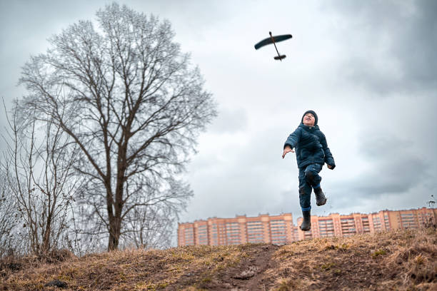 A boy is throwing toy airplane to the sky. He is dressed in casual clothes and rubber boots. The boy is jumping from the hillock and looking up at the flying homemade glider. Shooting at overcast spring day in a countryside near town