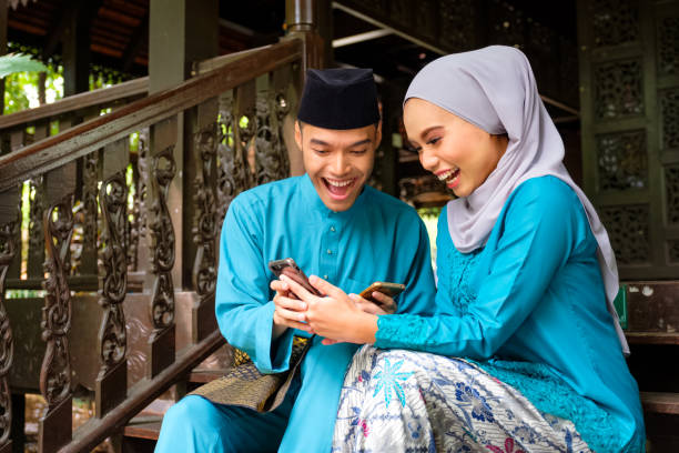 Young couple of malay muslim in traditional costume watching online content in a smart phone with happy expression during Eid al-Fitr celebration. Young couple of malay muslim in traditional costume watching online content in a smart phone with happy expression during Eid al-Fitr celebration at wooden stair. Raya and Muslim fashion concept. happy malay couple stock pictures, royalty-free photos & images