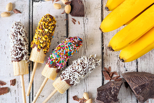 Frozen chocolate dipped banana pops with coconut, nuts and sprinkles. Above view table scene over a rustic white wood background.