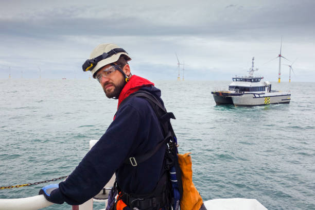 istock Serious technician waiting for transfer vessel on offshore platform - wind turbine and looking in camera. Offshore Wind farm is behind. 1219217179