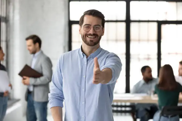 Smiling young Caucasian businessman stretch hand welcome new employee or worker at workplace, happy European male boss or CEO greeting meeting newcomer in office, employment concept