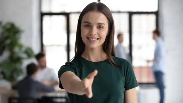 Headshot portrait of smiling young Caucasian businesswoman stretch hand welcome new employee at workplace, happy motivated female recruiter greeting meeting newcomer in office, employment concept