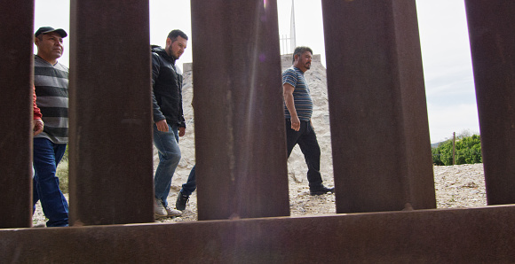 Several Hispanic Men Walk along the Mexican Side of the US/Mexican Steel-Slat Border Wall as the Camera Films on the US Side on a Sunny Day