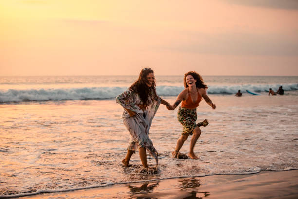 Serene Sunsets A wide shot of two female friends running out of the sea at Kuta beach in Bali, Indonesia at dusk while holding hands and laughing with each other. expatriate photos stock pictures, royalty-free photos & images