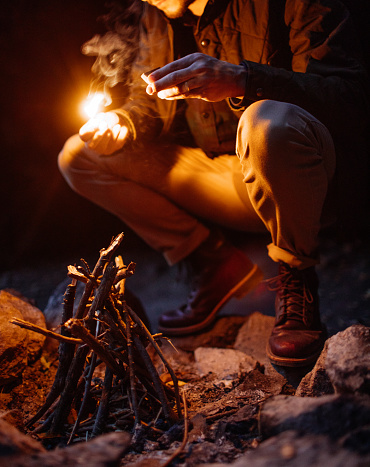 Man lighting a fire at dusk on a camping trip in New York. Glow from firelight on the man’s face as he lights fire in a ring of rocks.