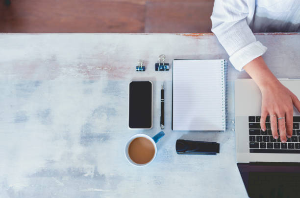 Top view of woman using laptop while sitting at the table with laptop, mobile phone, blank note pad and pen.
