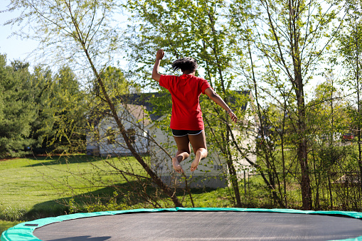 A shot of a young boy jumping up and down on a trampoline in the garden on a summers day, he is having fun and taking a selfie on a smartphone, his cousin is watching.
