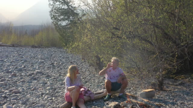 Mature couple prepare campfire on river shorelines, springtime