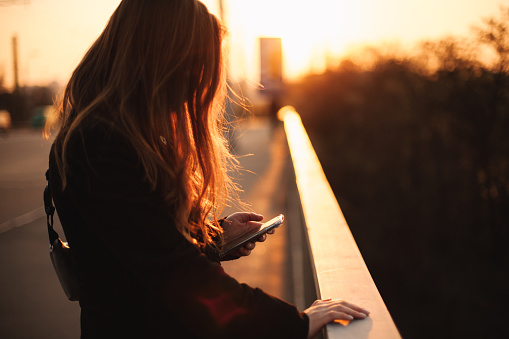 Young woman using smart phone while holding railing standing on bridge in city at sunset