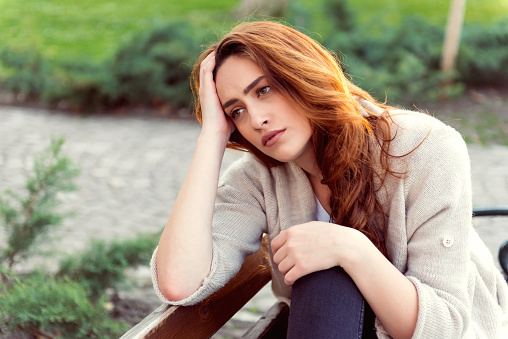 A girl with mahogany hair is sitting on a bench looking pensive. She is in a park and is dressed casually. She is resting her head on her hand. She might be sad or serious. With copy space.