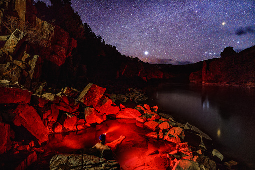 Man Soaking in Outdoor Natural Hot Springs Under the Stars - Night scenic view of stars and man soaking in natural hot spring along scenic river with very dark skies red light illuminating water and surrounding area.