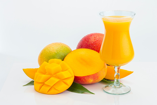 Mango composition on white background, two whole, one cut and a glass of mango juice. The cut slice has cubes pattern. There are also some pieces of mango and leaves of mango tree in the composition.