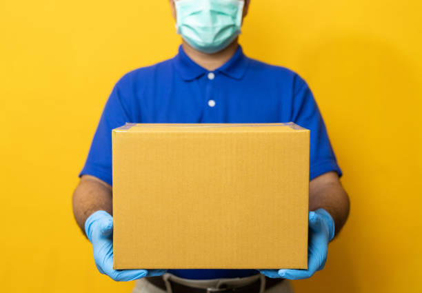 黄色の背景に小包の段ボール箱を保持しているゴム手袋とマスクを身に着けている配達員青い制服。 - men mail manual worker human hand ストックフォトと画像