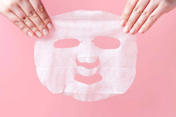 女性の手はピンクの背景に白い生地のマスクを保持しています。 - フェイスパック ストックフォトと画像