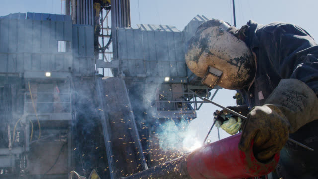 Slow Motion Shot of an Oilfield Worker Welding Two Pipes Together as Sparks Fly Next to a Derrick at an Oil and Gas Drilling Pad Site on a Sunny Day