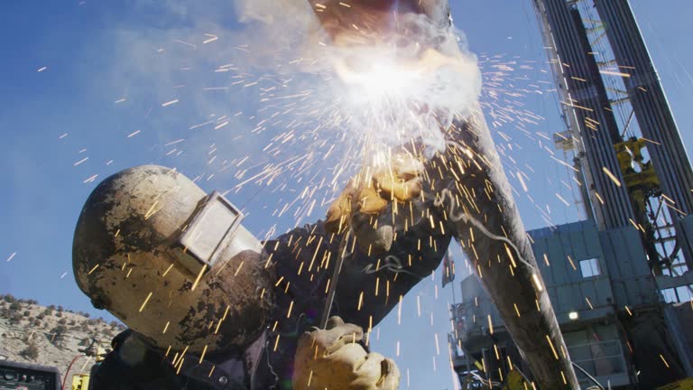 Slow Motion Shot of an Oilfield Worker Welding Two Pipes Together as Sparks Fly Next to a Derrick at an Oil and Gas Drilling Pad Site on a Sunny Day