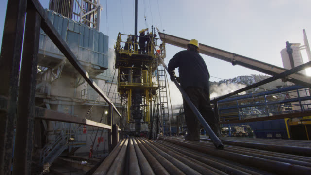 Slow Motion Shot of Oilfield Workers Rigging Up Drilling Pipe on a Tall, Metal Platform at an Oil and Gas Drilling Pad Site on a Cold, Sunny, Winter Morning