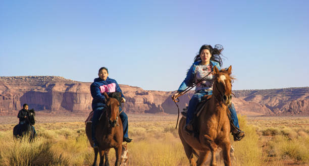 teenaged native american children (navajo) gallop on their horses through the monument valley desert in arizona/utah an einem sonnigen nachmittag - monument valley navajo mesa monument valley tribal park stock-fotos und bilder