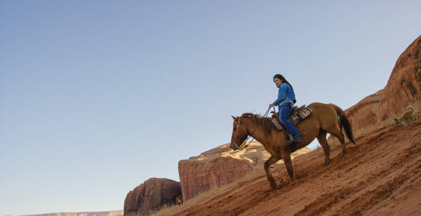 a teenaged native american girl (navajo) rides her horse down a steep hill with her dog in the monument valley desert in arizona/utah at sunset next to a large rock formation - monument valley navajo mesa monument valley tribal park stock-fotos und bilder
