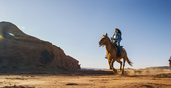 A Teenaged Native American Girl (Navajo) Rides Her Horse through the Monument Valley Desert in Arizona/Utah Next to a Large Rock Formation on a Clear, Bright Day