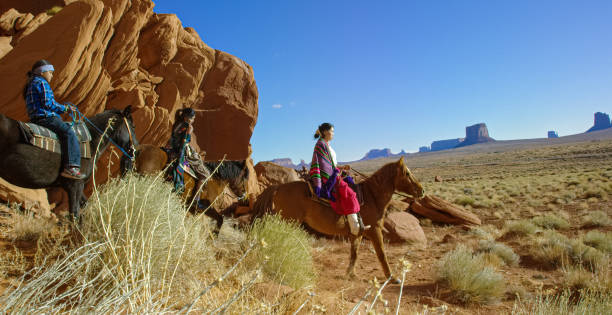 mehrere junge indianer (navajo) kinder, die traditionelle navajo-kleidung tragen, reiten ihre pferde durch die landschaft der monument valley wüste in arizona/utah neben einer großen felsformation an einem klaren, hellen tag - monument valley navajo mesa monument valley tribal park stock-fotos und bilder