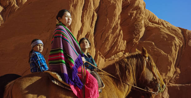 mehrere junge indianer (navajo) kinder tragen traditionelle navajo kleidung und sitzen auf ihren pferden und blicke auf die landschaft der monument valley wüste in arizona/utah neben einer großen felsformation - monument valley navajo mesa monument valley tribal park stock-fotos und bilder