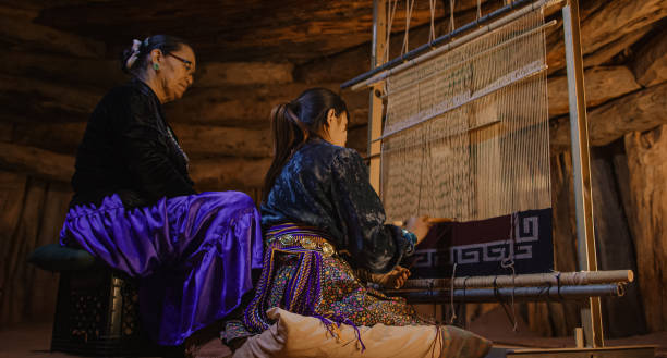 a native american grandmother (navajo) in her sixties teaches her teenaged granddaughter how to weave at a loom indoors in a hogan (navajo hut) - india traditional culture indigenous culture women imagens e fotografias de stock