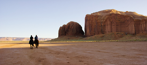 Two Native American Children (Navajo) Ride Their Horses in the Monument Valley Desert of Arizona/Utah Next to a Large Rock Formation at Sunset