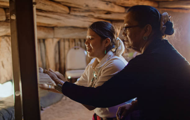A Native American Grandmother (Navajo) in Her Sixties Teaches Her Teenaged Granddaughter How to Weave at a Loom Indoors in a Hogan (Navajo Hut) A Native American Grandmother (Navajo) in Her Sixties Teaches Her Teenaged Granddaughter How to Weave at a Loom Indoors in a Hogan (Navajo Hut) loom photos stock pictures, royalty-free photos & images