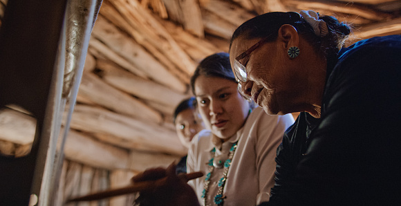 Una abuela nativa americana (Navajo) en sus sesenta enseña a sus nietas adolescentes cómo tejer en un telar en el interior de un Hogan (Navajo Hut) photo